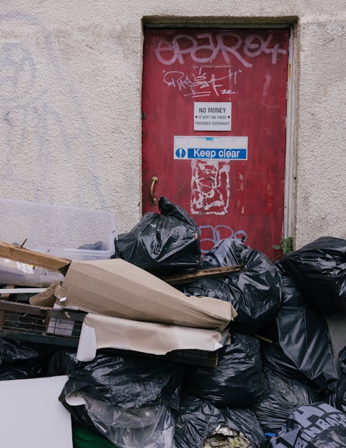 A black plastic rubbish bag and various discarded items including a flattened cardboard box and a white plastic object are piled loosely in front of a red metal door, which is set into a textured, off-white concrete wall. The door is covered with graffiti tags in pink and white spray paint, and features several signs, including a white one stating 'NO MONEY' with additional smaller text, and a blue sticker that reads 'Keep clear.' The surrounding environment appears to be an alleyway or side area, with the rubbish likely placed outside an entrance for collection or disposal, which suggests a scene of independent waste management. The lighting is natural, with no visible shadows, and the scene has an urban, utilitarian atmosphere associated with non-commercial waste handling. Waste Collection Islington specializes in rubbish removal, and this image visually supports the context of on-site clearance and private waste disposal methods often used as alternatives to council collection in the borough. The clutter and materials hint at a typical scenario of local rubbish accumulation awaiting collection.