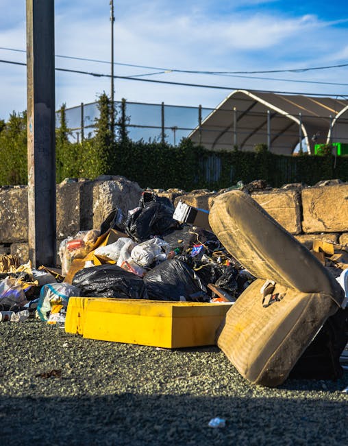 A roadside pile of waste and rubbish, including black plastic garbage bags, a yellow plastic container, and a worn, dirty car seat with a fabric cover, sits against a low stone wall. The rubble and waste are scattered across an asphalt surface, with the background featuring a metal lamppost, green hedges, and an open-air sports facility or arena with a curved roof and a dark fence. Overhead, the sky is partly cloudy with some blue visible, and electrical wires run horizontally across the scene, indicating an urban or suburban environment. The image illustrates an instance of improper rubbish disposal, which may be relevant to private waste collection or alternative rubbish removal services provided by Waste Collection Islington in comparison to municipal waste management options.