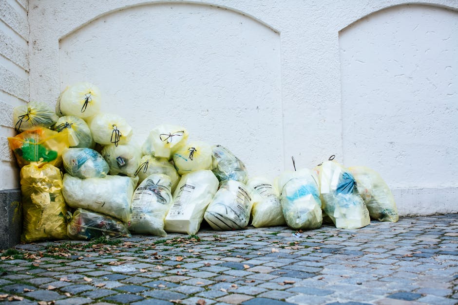 A collection of clear, yellow, and green plastic bags filled with trash, stacked against a white textured exterior wall of a building. The bags are tied with black twist ties and contain various types of waste, including paper, cardboard, and plastic items, with some bags appearing slightly bulging and others neatly closed. They are positioned on a cobblestone pavement that features small, irregularly shaped stones in shades of gray, brown, and black, with some scattered leaves and debris visible around the base of the bags. The scene suggests a nearby rubbish collection point, possibly managed by Waste Collection Islington, indicating an alternative waste handling method where bulk waste is temporarily stored outdoors awaiting removal. The natural daylight illuminates the scene, casting soft shadows, and emphasizing the textures of the bags, wall, and cobblestones, showcasing a typical urban waste disposal setup for private or independent rubbish collection services.
