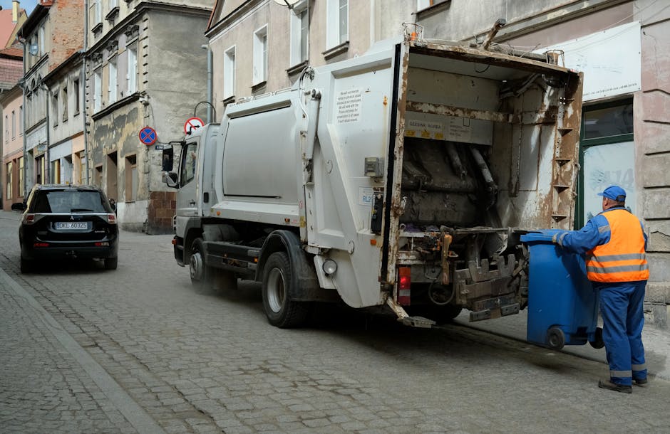 A worker in a high-visibility orange vest and blue uniform is seen emptying a blue wheeled trash bin into the rear compartment of a white commercial rubbish collection truck. The truck is parked on a narrow cobbled street surrounded by old, multi-story buildings with mixed brick and stucco facades. The collection compartment of the truck is open, revealing a rusted interior and mechanical components used for compacting waste. A small, dark grey car is parked nearby on the street, and there are no other vehicles or pedestrians visible. The scene is illuminated by natural daylight, highlighting the textures of the cobblestones, building surfaces, and the weathered metal of the truck. This imagery depicts an independent waste collection operation, typical for private rubbish removal services offered by companies like Waste Collection Islington, in an urban environment focused on non-local authority waste handling and on-site clearance efforts.