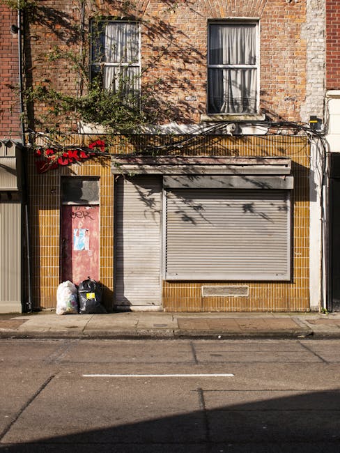 The image depicts the facade of a closed shop or commercial unit with a grey metal roller shutter partially down, situated beneath a row of two residential windows with white frames and dark curtains on the upper floor of a brick building. The brickwork is reddish-brown with some weathered areas, and a strip of yellow and brown tiles borders the storefront area, indicating an older or weathered exterior. To the left of the shutter, there is a small door with graffiti and peeling paint, partially obscured by shadows. In front of the building, on the pavement, there are three black and white plastic rubbish bags filled with waste, placed next to each other, with additional plastic bags and loose debris nearby. Above, a black cable runs horizontally across the facade, attached to the building, and an air conditioning unit is visible above the shutter, with shadows cast on the brick wall by nearby trees. The scene is illuminated by natural daylight, emphasizing the texture of the brickwork and the materials of the shopfront, which is consistent with a location where independent rubbish collection and private waste handling services such as those offered by Waste Collection Islington might operate in the area.