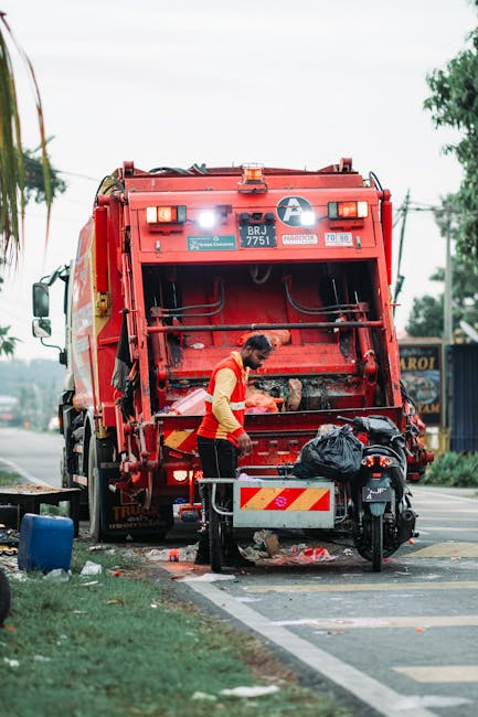 A worker in a high-visibility orange vest and blue uniform is seen emptying a blue wheeled trash bin into the rear compartment of a white commercial rubbish collection truck. The truck is parked on a narrow cobbled street surrounded by old, multi-story buildings with mixed brick and stucco facades. The collection compartment of the truck is open, revealing a rusted interior and mechanical components used for compacting waste. A small, dark grey car is parked nearby on the street, and there are no other vehicles or pedestrians visible. The scene is illuminated by natural daylight, highlighting the textures of the cobblestones, building surfaces, and the weathered metal of the truck. This imagery depicts an independent waste collection operation, typical for private rubbish removal services offered by companies like Waste Collection Islington, in an urban environment focused on non-local authority waste handling and on-site clearance efforts.