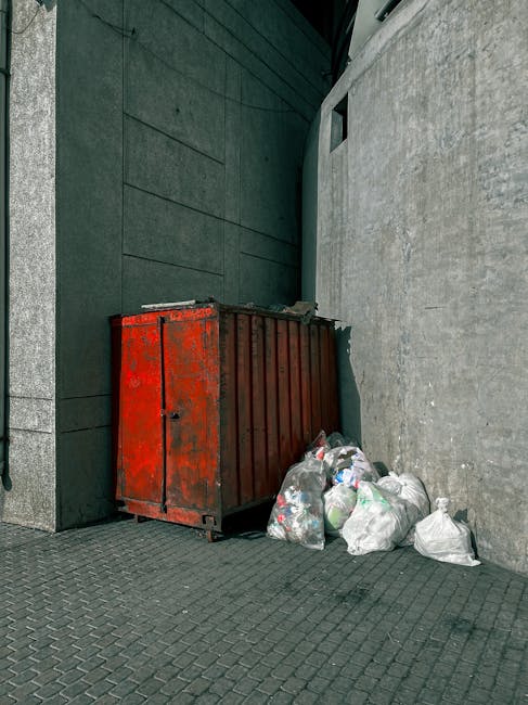A red metal skip with visible rust and chipped paint, positioned in a narrow urban alleyway against a dark green wall on the left and a light grey concrete wall on the right. The skip is partially filled with miscellaneous waste, and several white plastic rubbish bags are placed directly on the cobbled pavement in front of it, some slightly bulging or tied at the top. The environment suggests a backstreet or side access area typical for private waste disposal, with the concrete and metal surfaces showing signs of weathering. The overall scene appears calm and orderly, with no visible activity or people, reflecting an informal or alternative rubbish collection setup commonly used outside designated public refuse systems, aligning naturally with waste management services offered by companies like Waste Collection Islington.