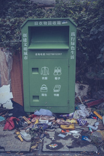 A red metal skip with visible rust and chipped paint, positioned in a narrow urban alleyway against a dark green wall on the left and a light grey concrete wall on the right. The skip is partially filled with miscellaneous waste, and several white plastic rubbish bags are placed directly on the cobbled pavement in front of it, some slightly bulging or tied at the top. The environment suggests a backstreet or side access area typical for private waste disposal, with the concrete and metal surfaces showing signs of weathering. The overall scene appears calm and orderly, with no visible activity or people, reflecting an informal or alternative rubbish collection setup commonly used outside designated public refuse systems, aligning naturally with waste management services offered by companies like Waste Collection Islington.
