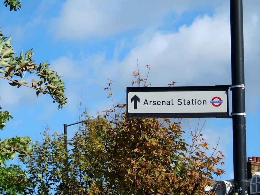 A rectangular street directional sign mounted on a black metal pole, pointing straight ahead towards 'Arsenal Station' with a black upward arrow symbol. The sign features the London Underground roundel logo on the right side, with the station name in black text on a white background. Surrounding the pole are leafy trees with green and brown foliage, some branches extending into the frame from the left and above, partially obscuring the sign. The background shows a bright blue sky with scattered white clouds. The scene suggests an urban environment, possibly near a residential or commercial area, where local rubbish collection services like Waste Collection Islington operate to facilitate waste management amid city infrastructure.