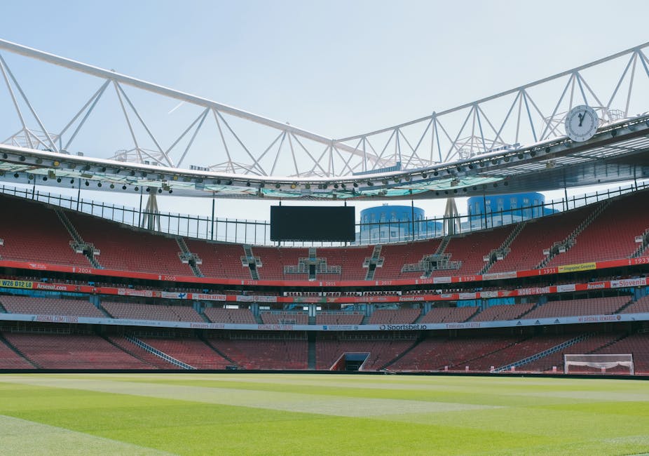 The interior of a soccer stadium featuring an empty grass pitch with well-maintained, lush green turf and clearly marked white boundary lines. Surrounding the pitch, there are tiered seating sections with predominantly red and grey seats, some sections having visible sponsorship banners in various colours. Above the seating areas, a large, rectangular electronic scoreboard is suspended, currently turned off, with a black screen. The stadium's partially covered roof structure consists of white, steel framework with a geometric truss design, supporting a translucent canopy that allows natural daylight to illuminate the interior. Large, circular clock faces are mounted on the roof's supporting beams. In the background, modern buildings with glass facades are visible beyond the stadium's upper tiers, contributing to the urban context. The overall scene appears bright and clear, emphasizing the open and spacious environment suitable for sporting events, and subtly aligning with potential private or independent event space management, as might be relevant to waste removal services supporting stadium maintenance.