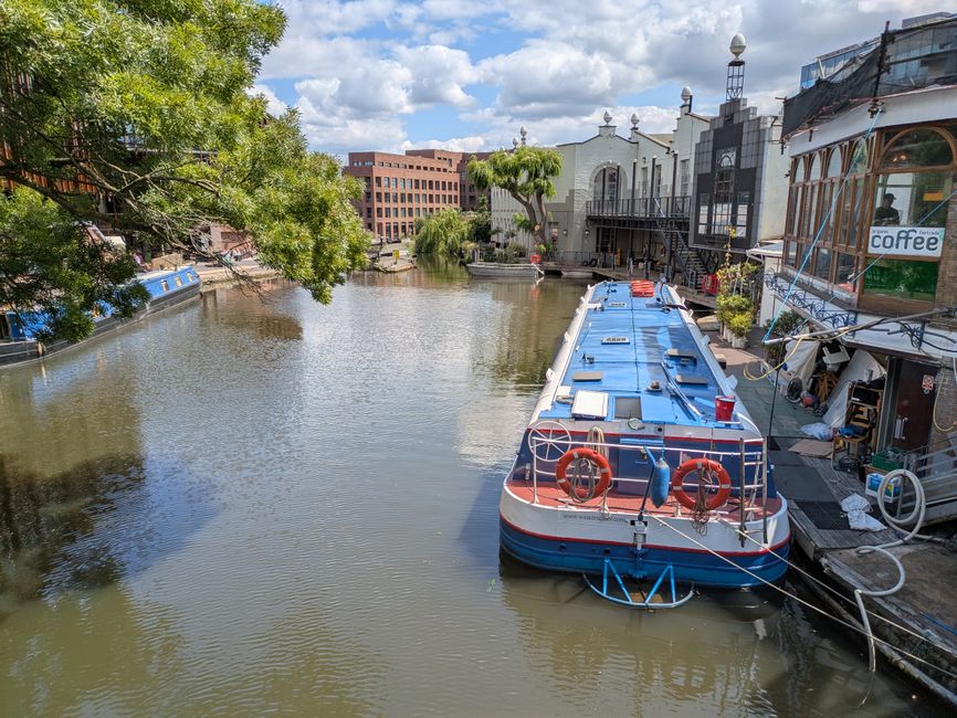 A narrow waterway running through a lush, green urban area on a bright, partly cloudy day. The canal's calm surface reflects patches of the blue sky and scattered white clouds. On the right side, a paved walkway runs parallel to the water, with several pedestrians walking along it, some pushing strollers. Parked cars line the edge of the street adjacent to the path, with a few small boats moored along the water's edge, some covered with protective tarps. The left side of the canal is bordered by dense trees and shrubs, creating a natural, shaded environment, while on the right, there are residential buildings with brick facades and large windows, some partially obscured by leafy tree canopies. In the background, a church spire rises above the treetops, and additional urban structures are visible further along the waterway. The scene illustrates a typical waterside setting, where private and community activities such as walking and boating take place, which could relate to local rubbish removal or waste management efforts by Waste Collection Islington serving this waterside community.