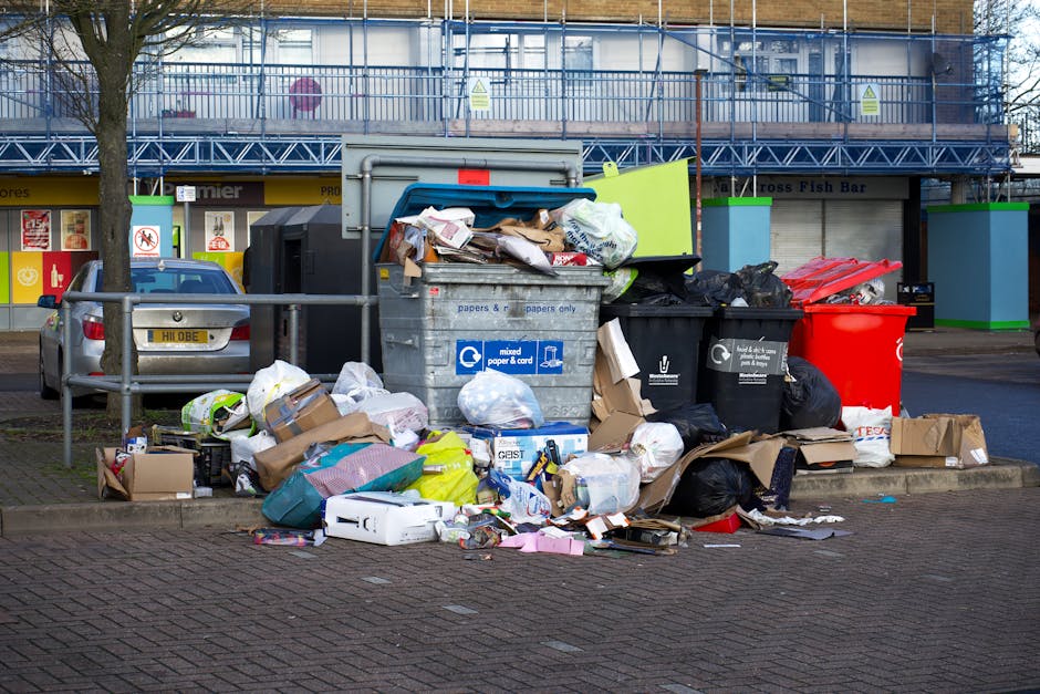 A large collection of overflowing rubbish bins and scattered waste packaging on a paved sidewalk in front of a commercial building in an urban area. The piles consist of black, red, and grey bins, some with lids open, revealing mixed paper, cardboard, plastic bags, and miscellaneous rubbish. Cardboard boxes and paper packaging are strewn around at the base of the bins, with some flattened and others crumpled. In the background, there is a building with multiple store fronts, including a fish bar, and an upper floor covered in scaffolding and protective netting, indicating ongoing construction or renovation. To the left, a parked grey vehicle is partially visible behind a metal railing, which separates the waste area from the parked car. The scene is evenly lit with natural daylight, emphasizing the disorganized state of waste accumulation, which could be linked to private waste collection services or non-standard rubbish disposal methods outside regular council clearance processes, aligning with services offered by Waste Collection Islington in facilitating alternative waste handling solutions.